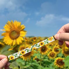 two hands holding a beaded bracelet in front of a sunflower field with blue sky