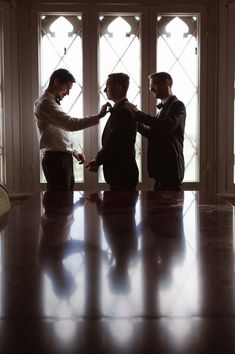 three men standing in front of a window with ties on their hands and looking at each other