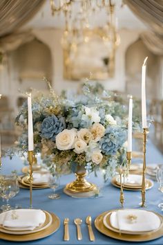 a table set with blue linens and gold place settings, white flowers and candles