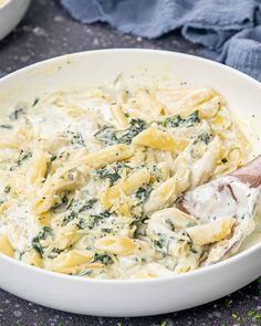 a white bowl filled with pasta and spinach on top of a blue cloth next to a wooden spoon