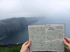 someone holding up an open book in front of a cliff overlooking the ocean on a cloudy day