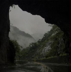 an empty road in the middle of a mountain with rain coming down and trees growing on both sides