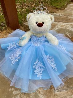 a teddy bear wearing a blue dress and tiara sitting on the ground next to a wooden post