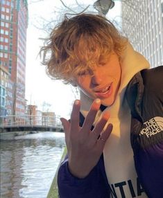 a young man with curly hair wearing a jacket and holding his hand up to the camera