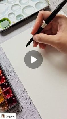 a person holding a pen and writing on a piece of paper with watercolors in the background