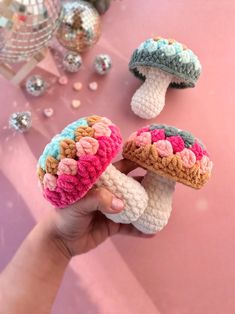 crocheted mushrooms are being held up by someone's hand on a pink surface