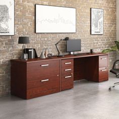 an office desk with two computer monitors and a printer on it in front of a brick wall