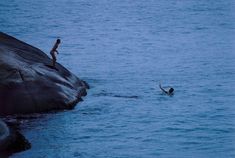 two people are swimming in the water near some large rocks and one person is jumping into the water