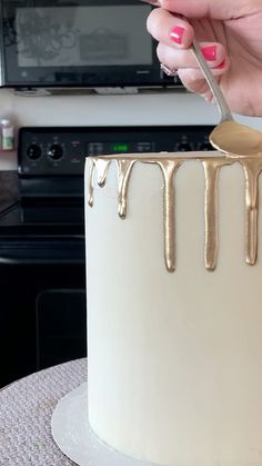 a woman is decorating a white cake with gold icing on it and holding a spoon