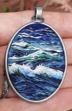 a hand holding an oval pendant with blue and green waves on the ocean in it