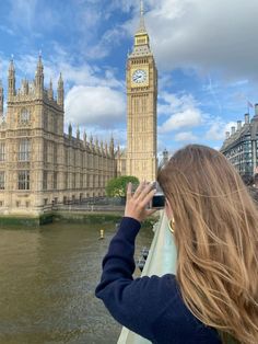 a woman taking a photo of the big ben clock tower from across the river thames