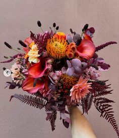 a hand holding a bouquet of flowers against a pink wall with ferns and other foliage