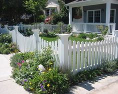 a white picket fence in front of a house with flowers and plants growing around it