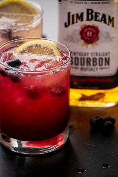 two glasses filled with red liquid next to a bottle of booze and lemon wedges