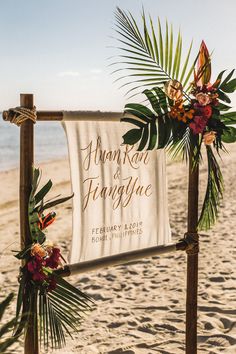 a sign on the beach with flowers and greenery hanging from it's posts