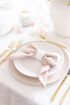 a table set with white plates and pink napkins, gold forks and silverware