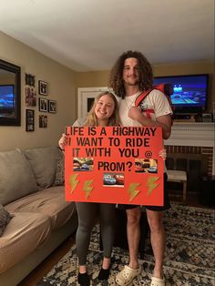 a man and woman standing in front of a couch holding a sign that says life is a highway, i want to ride it with you from?