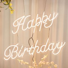 a happy birthday neon sign with flowers in the foreground and curtains in the background