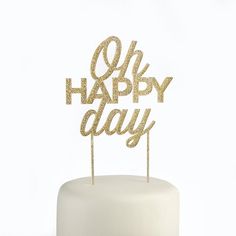 a white cake topped with a gold happy birthday cake topper and the words oh happy day spelled in cursive letters