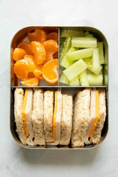 an open lunch box with sandwiches, orange slices and cucumbers in it on a white surface