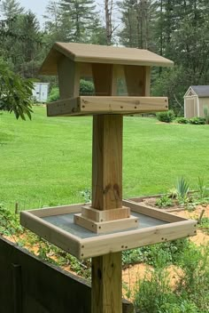 a wooden bird feeder in the middle of a yard with grass and trees behind it