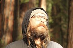 a man with long hair wearing glasses and a hoodie in the middle of a forest