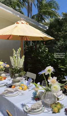 an outdoor dining table set with plates, flowers and fruit under an umbrella on a sunny day