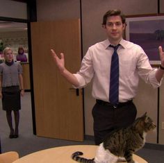 a man standing in an office with a cat on his lap and two people behind him