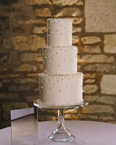 a three tiered cake with white frosting on a glass stand next to a brick wall