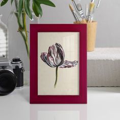 a red frame with a flower in it next to a camera and some pens on a table