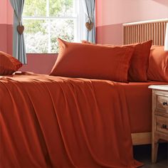 a bed with orange sheets and pillows next to a window in a pink walled room