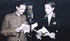 two men standing next to each other in front of a microphone holding papers and looking at something