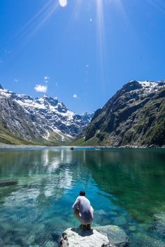 a man sitting on top of a rock in the middle of a lake surrounded by mountains