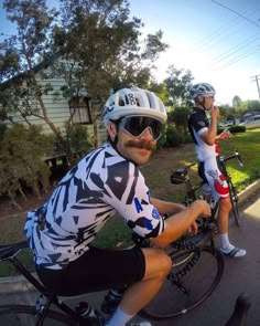 two bicyclists are riding down the street with their helmets on and one has a moustache