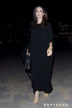 a woman is walking down the street at night wearing a long black dress and carrying a handbag