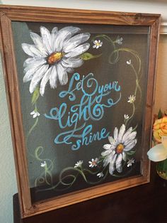 a painting that says let your light shine with daisies in front of it on a table