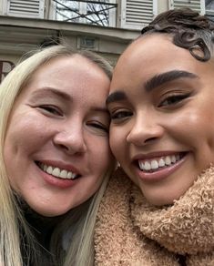 two women smiling and hugging each other in front of a building with white shutters