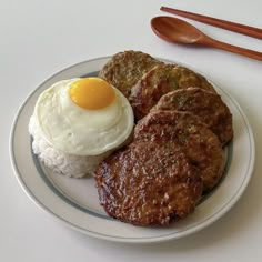 a white plate topped with meat, rice and an egg next to chopsticks