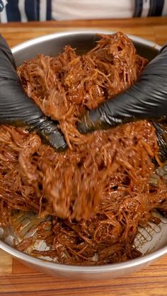 two hands with black gloves are in a silver bowl filled with shredded meat and vegetables