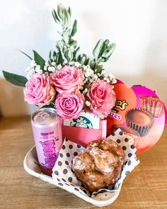 a basket filled with donuts and pink flowers on top of a wooden table next to a can of soda