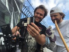 two men standing next to each other while holding up a camera and a wooden stick