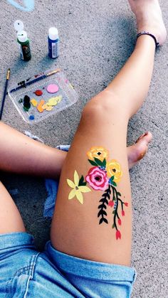 a woman with flowers painted on her stomach sitting next to paintbrushes and paints