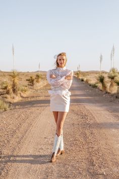 a woman is standing in the middle of a dirt road wearing boots and a white dress