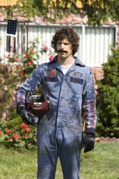 a man in blue overalls holding a red and white helmet on the grass with flowers behind him