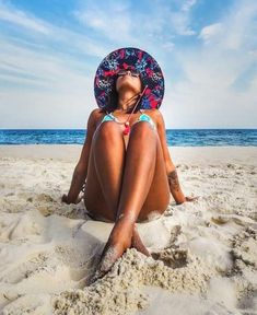 a woman sitting in the sand at the beach wearing a sunhat on her head