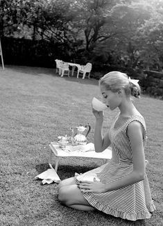 a woman sitting on the grass drinking from a teapot and holding a cup in her hand