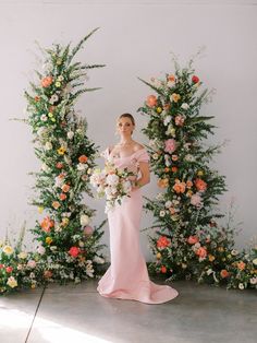 a woman in a pink dress standing under an arch with flowers and greenery on it