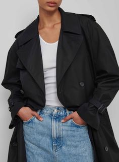 a woman wearing a black trench coat and white top is standing with her hands on her hips