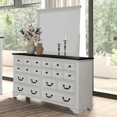 a white dresser and mirror in a room with large windows, flowers and candles on it