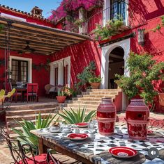 an outdoor dining table with red vases and plates on it in front of a house